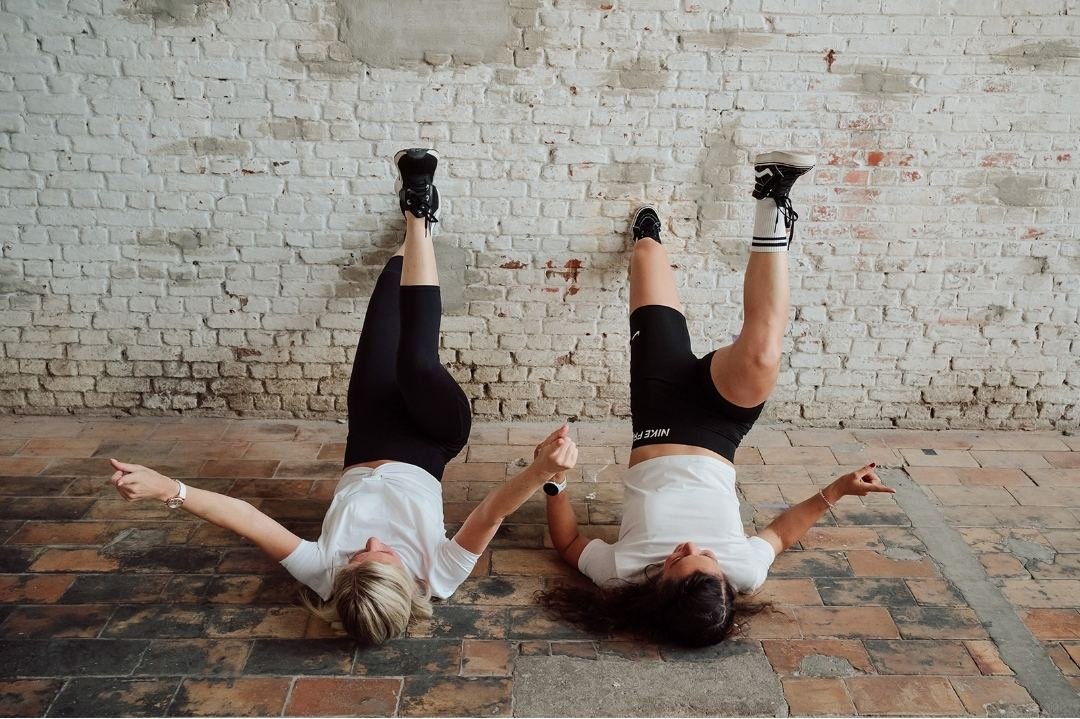 Twee sporters doen samen een oefening op de grond, met de benen tegen de muur